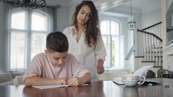 Jovem mãe e filho à mesa. Boy Writes In Notebook. Ele faz seu dever de casa. — Vídeo de Stock