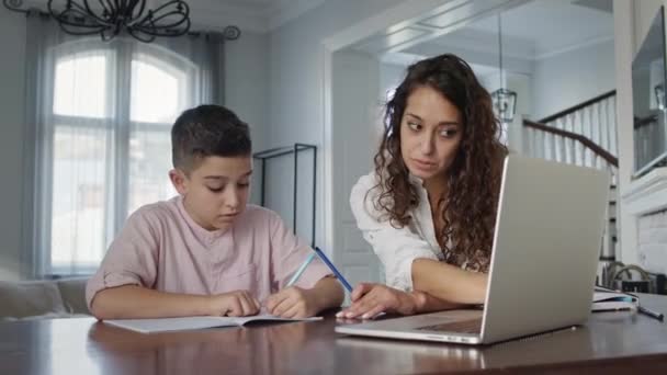 Junge Mutter und Sohn am Tisch. Junge schreibt in Notizbuch. Er macht seine Hausaufgaben. — Stockvideo