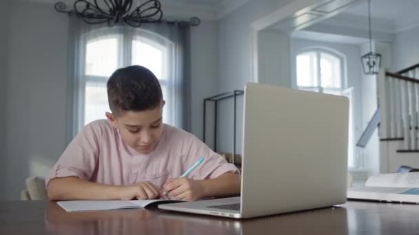 Junge von acht Jahren sitzt vor Laptop am Tisch. Er schreibt in Notizbuch. Junge macht Hausaufgaben. — Stockvideo