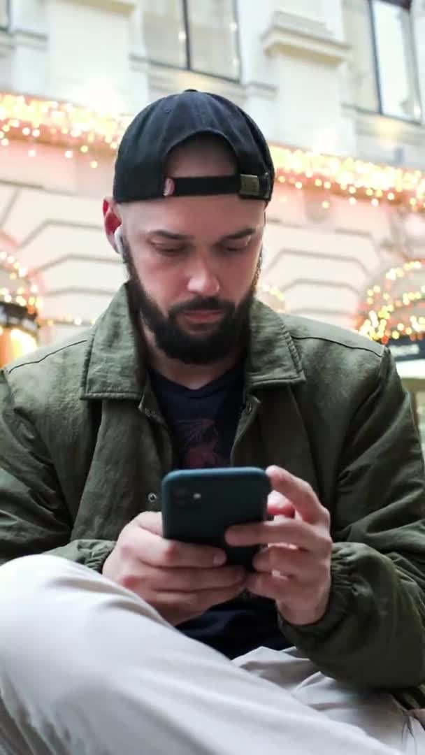 Mladý muž v čepici s plnovousem sedí na ulici s telefonem ve svých rukou. Pozvedne obočí v překvapení a úsměvu. — Stock video