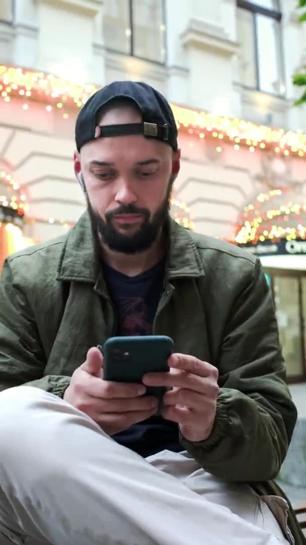 Jongeman met baard op straat zittend met telefoon in zijn handen. — Stockvideo