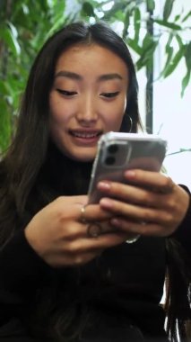 Elinde cep telefonuyla pencerenin kenarında oturan Koreli kız. Kız telefon ekranına bakıyor. Kapat..