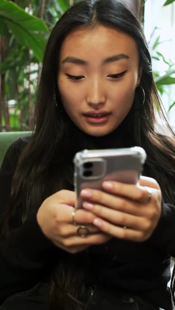 Koreanska flicka sitter vid fönstret med mobiltelefon i händerna. Flickan tittar på telefonskärmen och skrattar. Närbild. — Stockvideo