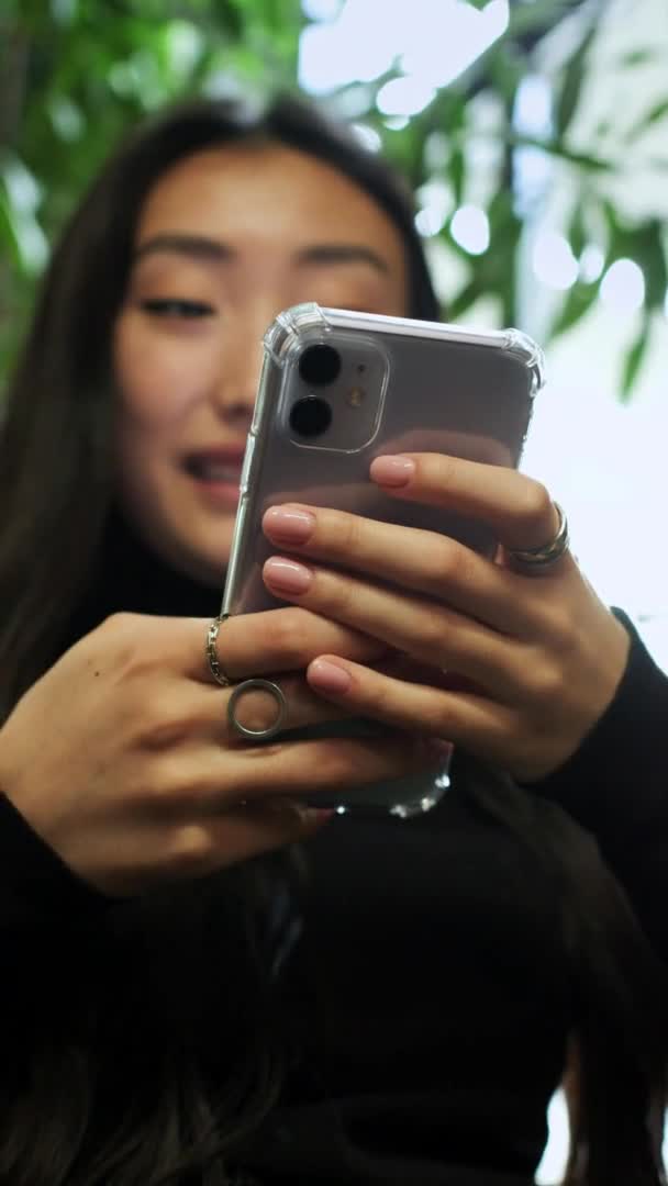 Koreanska flicka sitter vid fönstret med mobiltelefon i händerna. Flickan tittar på telefonskärmen och skrattar. — Stockvideo