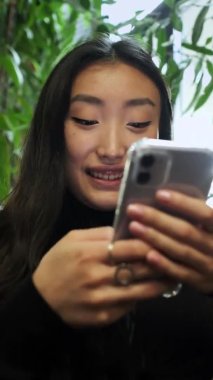 Elinde cep telefonuyla pencerenin kenarında oturan Koreli kız. Kız telefon ekranına bakıyor. Kapat..