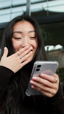Elinde cep telefonu olan Koreli bir kızın portresi. Kız telefon ekranına bakıyor. Yakın çekim. Kız eliyle ağzını kapatıyor..