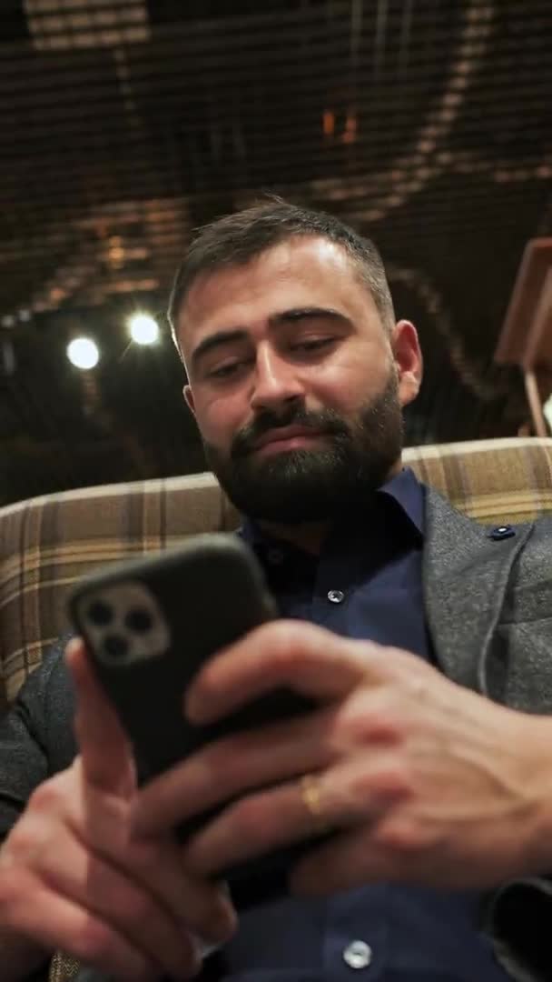 Homem bonito senta-se no sofá com telefone em suas mãos. homem elegante de cabelos escuros com barba usa o telefone. — Vídeo de Stock