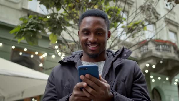 Молодой черный парень стоит на улице с телефоном в руках. Он смотрит на телефон и улыбается. — стоковое видео