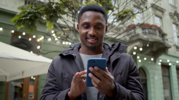 Mladý černoch stojí na ulici s telefonem v ruce. Dívá se na telefon a usmívá se. — Stock video