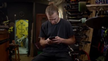 Elinde telefonu olan genç adam bisiklet atölyesinde oturuyor. Aktif olarak telefonu kullanıyor..