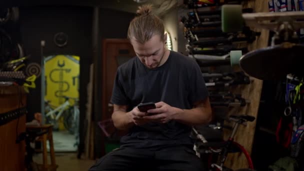 Jongeman met telefoon in zijn handen zittend in de fietsenwinkel. Hij gebruikt actief de telefoon.. — Stockvideo