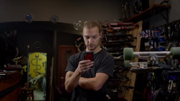 Junger Mann mit Handy in der Hand steht in Fahrradwerkstatt Er nutzt das Telefon aktiv. — Stockvideo