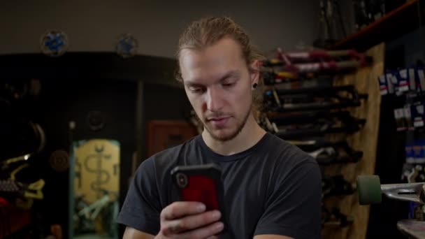 Mladý muž s telefonem v ruce stojící v cyklistické dílně. Aktivně používá ten telefon. On zvedne obočí v překvapení. — Stock video