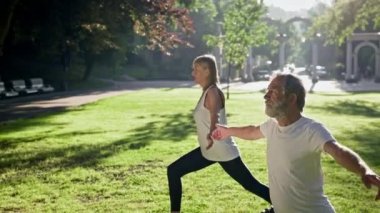 Yaşlı adam ve gri saçlı kadın parkta spor yapıyor. Bir ayağı önde durur ve avuçlarına dokunarak ellerini kaldırırlar. Güneş ışınları onları aydınlatır. Yaz Güneşli Günü.