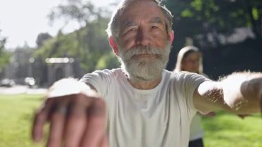 Park 'taki yaşlılar spor için içeri giriyor. Yaşlı, gri saçlı bir adam ve kolları açık bir kadın çömelmiş. Kocasının Arkasında Kadın. Güneşli Yaz Günü.