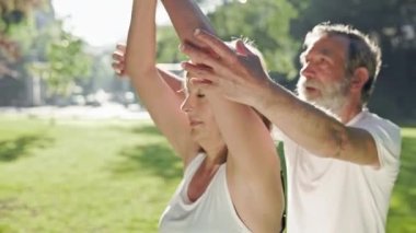 Yaşlı adam ve gri saçlı kadın parkta spor yapıyor. Ellerini kaldırır ve indirirler. Güneşli bir yaz günü. Erkek, Kadın Ellerini Düzeltti.
