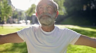 Yaşlı gri saçlı adam parkta spor yapıyor. Ellerini kaldırır ve indirir. Güneşli Yaz Günü.