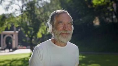 Yaşlı adam kulaklıklarla parkta koşuyor. Güneşli yaz gününde sabah koşusu. Sakallı Gri Adam.