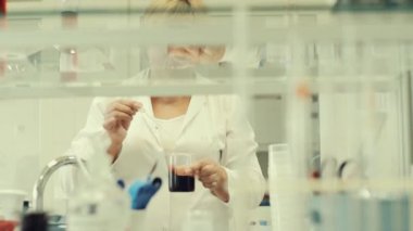 Laboratuvar teknisyeni sıvının içine reaktörü damlatıyor. Laboratuvar teknisyeni sıvıyı sallıyor. Siyah Sıvı büyük bir cam şişede. Beyaz bornoz giyen kadın..