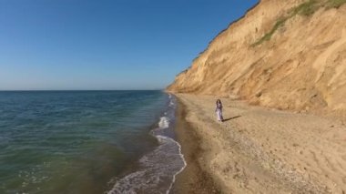 Güzel kız sahilde oynuyor ve dans ediyor. Çakıl taşlarında yalınayak koşan genç bir kadın. Uzun, açık renkli, bedava bir elbise moda. Esmer Kız Dönüyor.