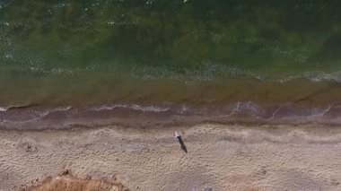 Hava görüntüsü. Sıcak bir plajda yürüyen genç bir kadın. Kızın gölgesi kumun üzerine düşüyor. Denizin Sakin Dalgaları. Model üzerinde uzun, açık, bedava bir elbise var.