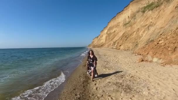 空中展望 美しい若い女性がビーチを歩いています 魅力的な女の子は小石の上に裸足で歩く ジプシー カクテル マキシドレスがモデルです 風がドレスを拾う — ストック動画