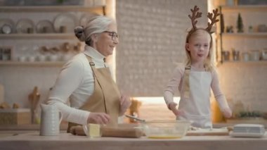 Noel tatilleri. Büyükannem ve küçük kızım mutfakta. Önlüklü yeni yıl pastası hazırlıyorlar. Eğleniyorlar ve dans ediyorlar. Kızın kafasında geyik boynuzu şeklinde sarı saç ve süslemeler var..