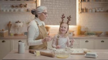 Noel tatilleri. Büyükannem ve küçük kızım mutfakta. Önlüklü yeni yıl pastası hazırlıyorlar. Kızın kafasında geyik boynuzu şeklinde sarı saç ve süslemeler var..