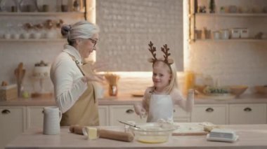 Noel tatilleri. Büyükannem ve küçük kızım mutfakta. Önlüklü yeni yıl pastası hazırlıyorlar. Kızın kafasında geyik boynuzu şeklinde sarı saç ve süslemeler var..