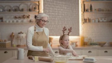 Noel tatilleri. Büyükannem ve küçük kızım mutfakta. Önlüklü yeni yıl pastası hazırlıyorlar. Eğleniyorlar ve dans ediyorlar. Kızın kafasında geyik boynuzu şeklinde sarı saç ve süslemeler var..