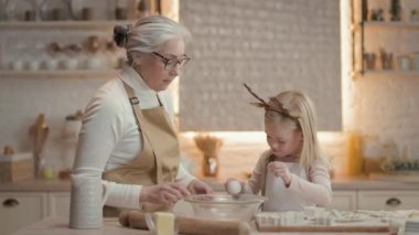 Büyükannem ve küçük kızım mutfakta. Arkalarında teçhizatlı bir sürü raf var. Kiz kase uzerine yumurta atip kiriyor. Büyükanne Spurs ve Kıza Yardım Ediyor. Noel Fırını Hazırlıyorlar.