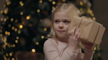 Hediye Kutusu Tutan Kız ve Taşıma. İçinde ne olduğunu dinlerdi. Sarışın, uzun saçlı, dört yıllık küçük kız. Arkasında Noel ağacı, çelenklerle parıldıyor.