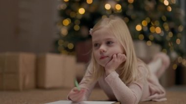 Sarışın, uzun saçlı, dört yıllık küçük kız. Arkasında Noel ağacı çelenklerle parıldıyor. Kız çizer. Noel ağacının yanında, yerde. Küçük Yastıklı Kız. Noel Ağacı Hediyelerinin Yakınları.