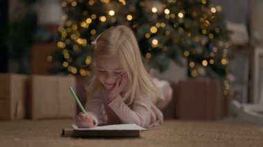 Sarışın, uzun saçlı, dört yıllık küçük kız. Arkasında Noel ağacı çelenklerle parıldıyor. Kız çizer. Noel ağacının yanında, yerde. Küçük Yastıklı Kız. Noel Ağacı Hediyelerinin Yakınları.