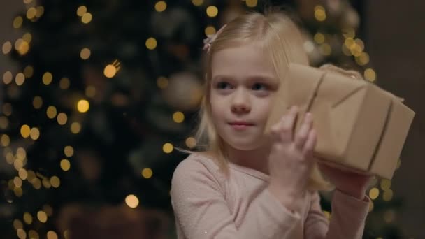 女の子はギフトボックスを保持し それを移動 彼女はそれの中のものを聞く ブロンドロングヘアで4年の女の子 背後にあるクリスマスツリー ガーランドの輝き — ストック動画
