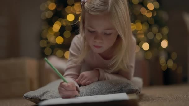 ブロンドロングヘアで4年の女の子 背後にはガーランドのクリスマスツリーが輝きます 女の子が描く 彼女はクリスマスツリーの近くの床にいる 枕の上の女の子 — ストック動画