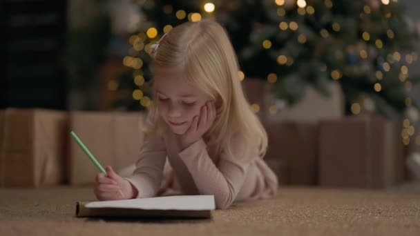 ブロンドロングヘアで4年の女の子 背後にはガーランドのクリスマスツリーが輝きます 女の子が描く 彼女はクリスマスツリーの近くの床にいる 枕の上の少女 クリスマスツリーギフトの近く — ストック動画
