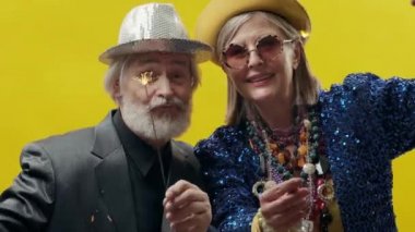 Yaşlı adam ve kadın. Sarı arka planda şık, yaşlı bir kadın. Şapka ve gözlük takıyor. Şapkalı, şık sakallı adam. Havai fişekleri olan şık bir çift..