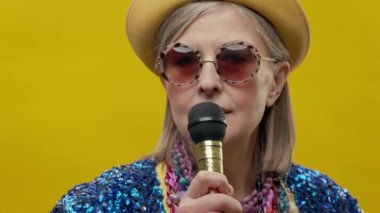 Sarı arka planda şık, yaşlı bir kadın. Şapka ve gözlük takıyor. Gülümser ve mikrofona şarkı söyler..