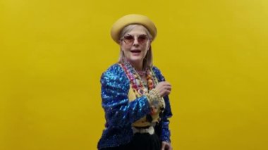 Sarı arka planda şık, yaşlı bir kadın. Şapka ve gözlük takıyor. Neşeli Kadın Dans Ediyor.