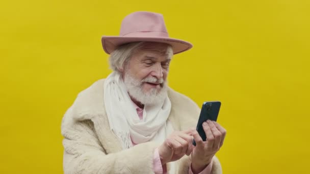 黄色の背景にピンクの服を着たスタイリッシュな老人 彼は髭と口ひげを生やして灰色です 帽子を頭にかぶっている 彼はそれを楽しみながら電話を続けます — ストック動画