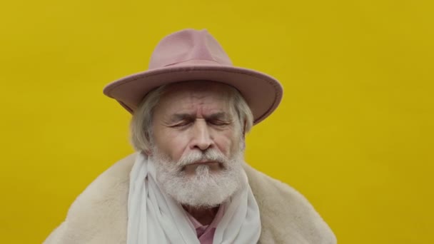 穿着粉色衣服的古板老人背景是黄色的 他是灰色与胡子和胡子 他头上有帽子 人的眨眼改变了他的表情 — 图库视频影像