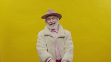Sarı Arkaplanda Pembe Giysiler Giyen Şık Yaşlı Adam. Gri sakallı ve bıyıklı. Kafasında şapkası var..