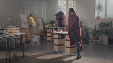 Stüdyoda Duran Genç Kız. Dergiye göz atıyor. Kısa saçlı, uzun ceketli hoş bir kadın. Odada bir sürü resim ve kolaj var. Yaratıcı Stüdyo. Elbiseli ve Dikişli Manken