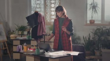 Stüdyoda çalışan genç kız moda tasarımcısı. Masada dizüstü bilgisayar ve çizimlerle duruyor. Onun kaleminde. İç mekan mankenleri ve kıyafetleri. Kısa saçlı, uzun ceketli tatlı kız..