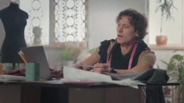 Masada Erkek Tasarımcı. Laptopa bakmaktan bahsediyor. Boynunda atkı ve santimetre şeridi var. Masadaki çizimler. Çalışma Akışı.