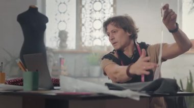 Masada Erkek Tasarımcı. Laptopa bakmaktan bahsediyor. Boynunda atkı ve santimetre şeridi var. Masadaki çizimler. Çalışma Akışı.