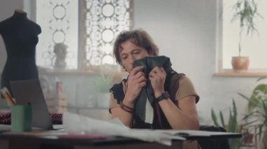 Masada Erkek Tasarımcı. Laptopa bakmaktan bahsediyor. Boynunda atkı ve santimetre şeridi var. Masadaki çizimler. İş akışı. Kumaşı Gösteriyor.