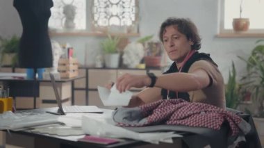 Masada Erkek Tasarımcı. Laptopa bakmaktan bahsediyor. Boynunda atkı ve santimetre şeridi var. Masadaki çizimler. Çalışma Akışı.