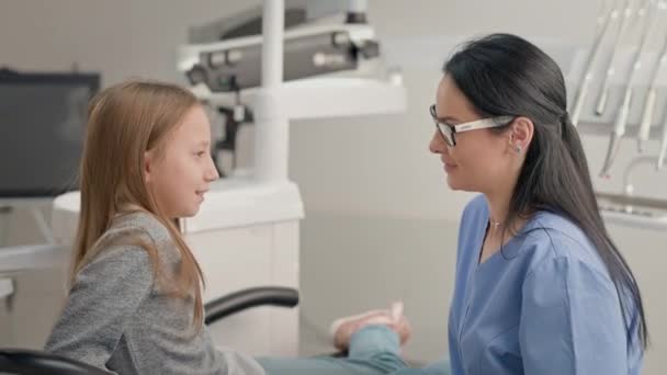 歯医者事務所の少女 歯科治療の前に女の子と話す若い女性医師 女の子は椅子に座っている 長髪のかわいい女の子は慎重に聞く — ストック動画